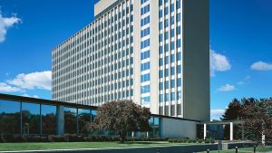 A low angle photo of the Admin 101 Building on NIST's Gaithersburg, Maryland campus. The building is tall with many windows, and the sky in the background is blue with few clouds.